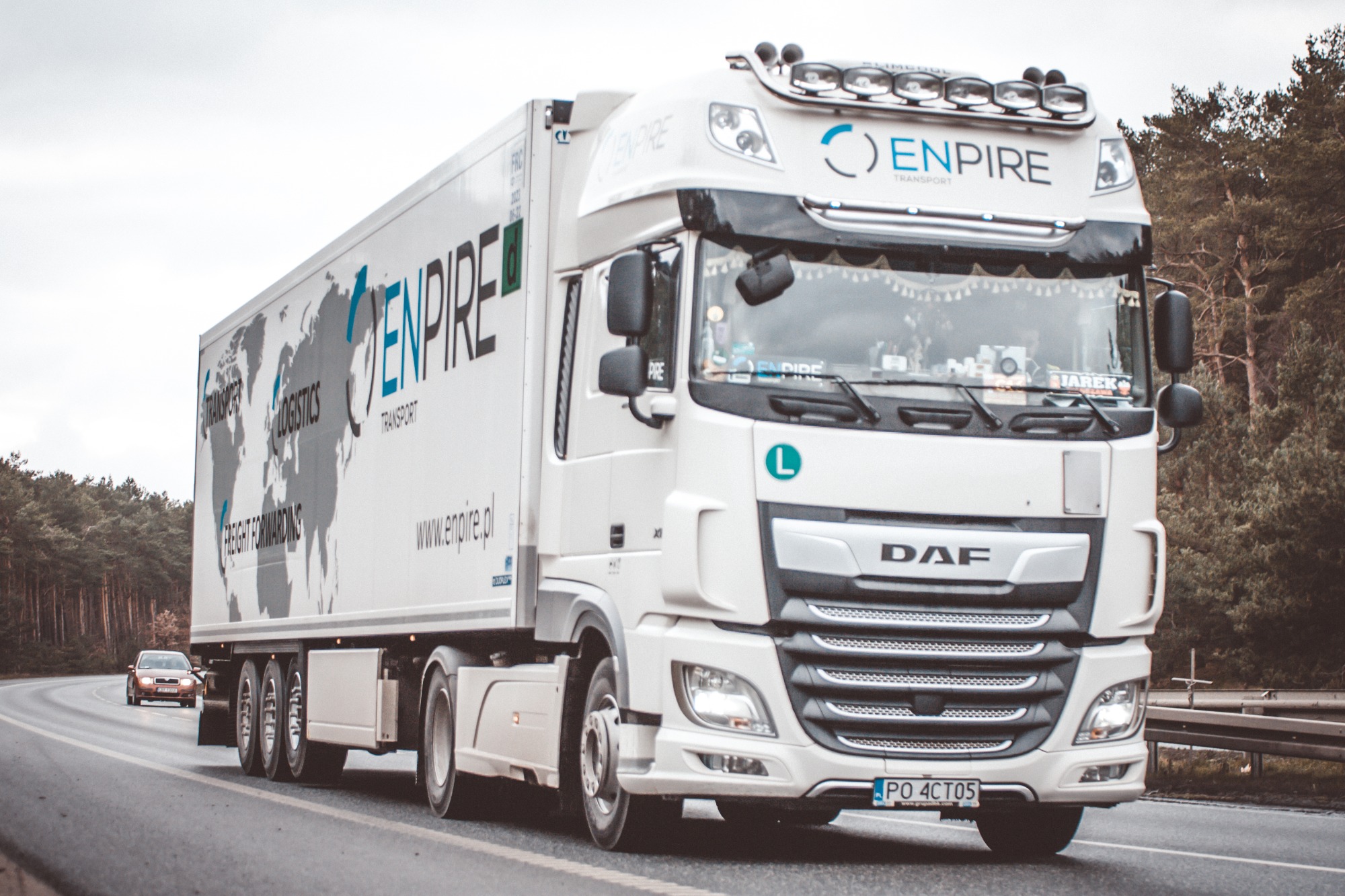Biała ciężarówka DAF z naczepą, ozdobiona mapą świata i logo firmy Enpire Transport, jadąca po asfaltowej drodze w kierunku widza, z lasem w tle pod pochmurnym niebem.