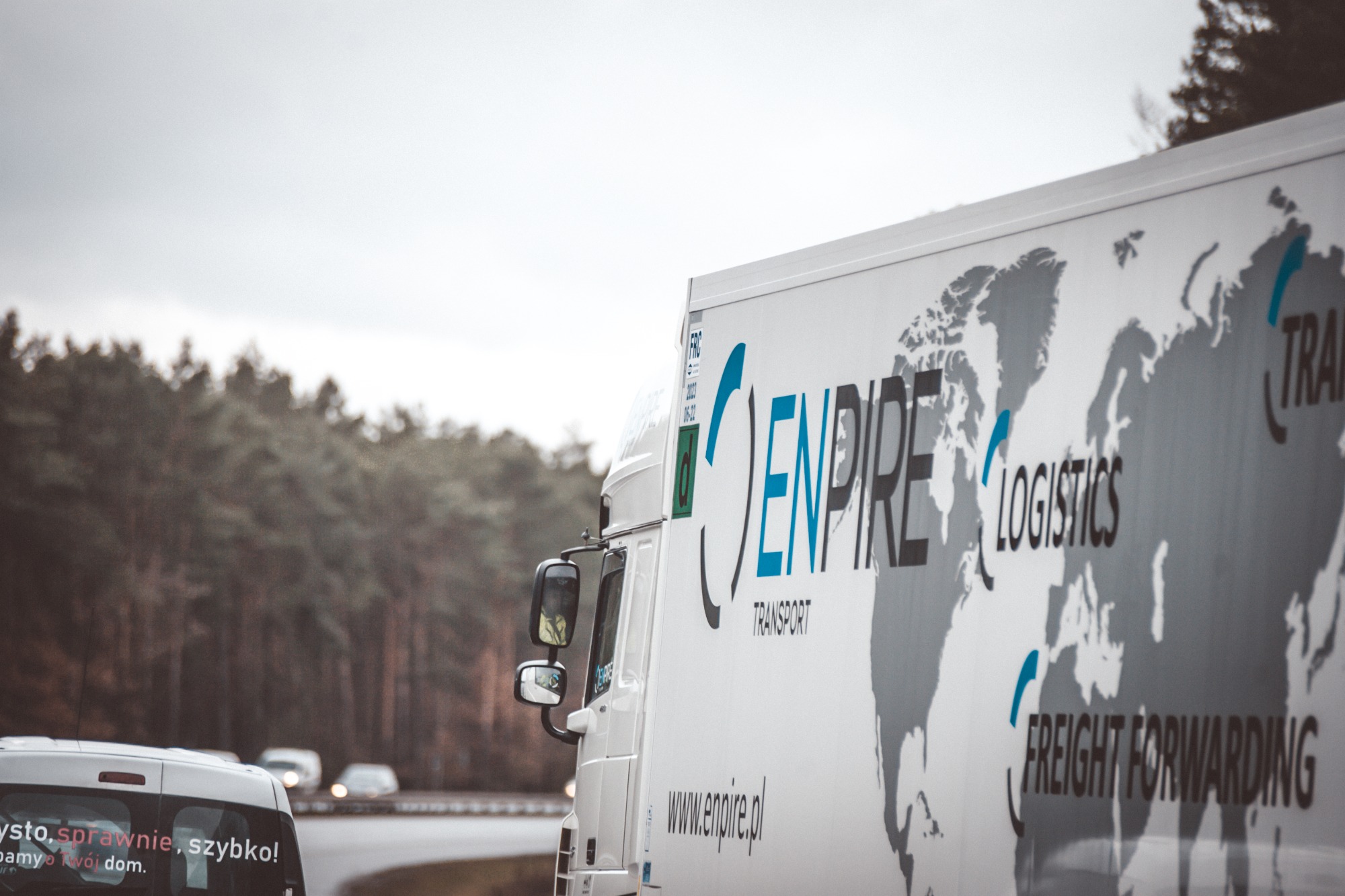 Biała ciężarówka z logo Enpire Logistics i mapą świata na naczepie, widoczna na drodze z zalesionym tłem w pochmurny dzień.