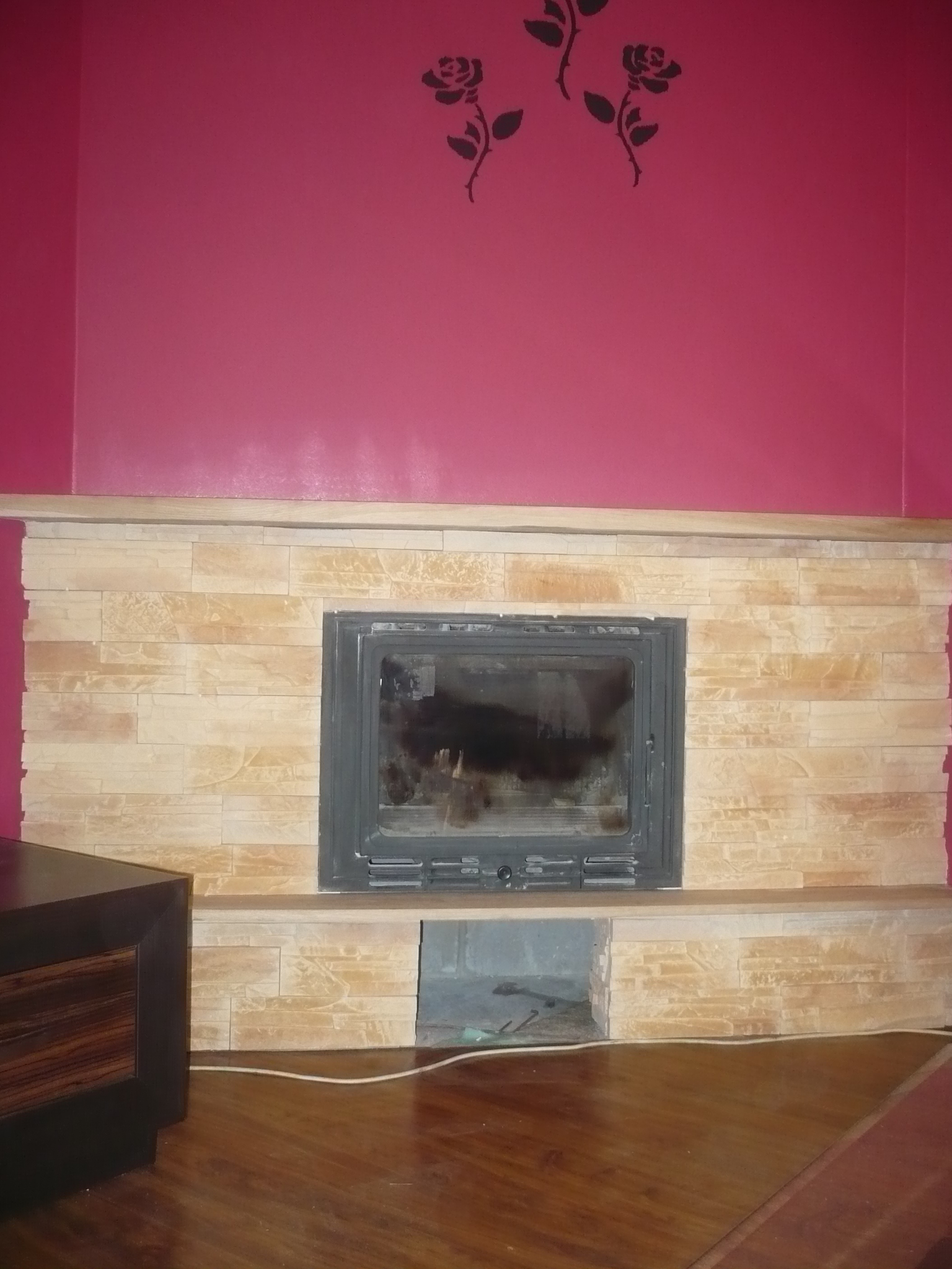 Obudowa kominka wykończona płytkami imitującymi kamień, z czarnym wkładem kominkowym, na tle różowej ściany z naklejkami w kształcie róż. Widoczny fragment drewnianej podłogi.