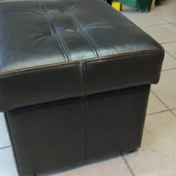 Pufa tapicerowana ciemną skórą z pikowaniem na siedzisku, widoczna na kafelkowej podłodze.