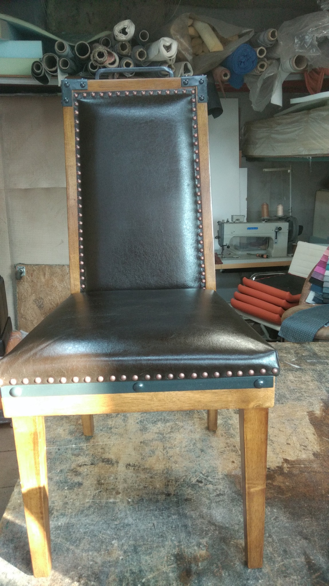 Krzesło po renowacji, obite brązową skórą z ozdobnymi nitami, widoczne narzędzia i materiały w tle, warsztat.