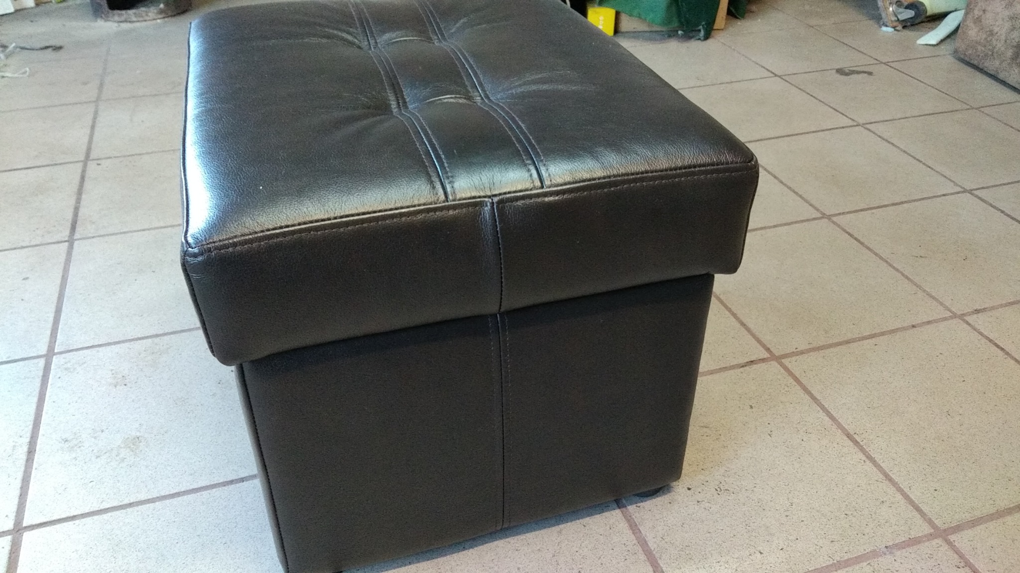 Pufa tapicerowana ciemną skórą z pikowaniem na siedzisku, widoczna na kafelkowej podłodze.