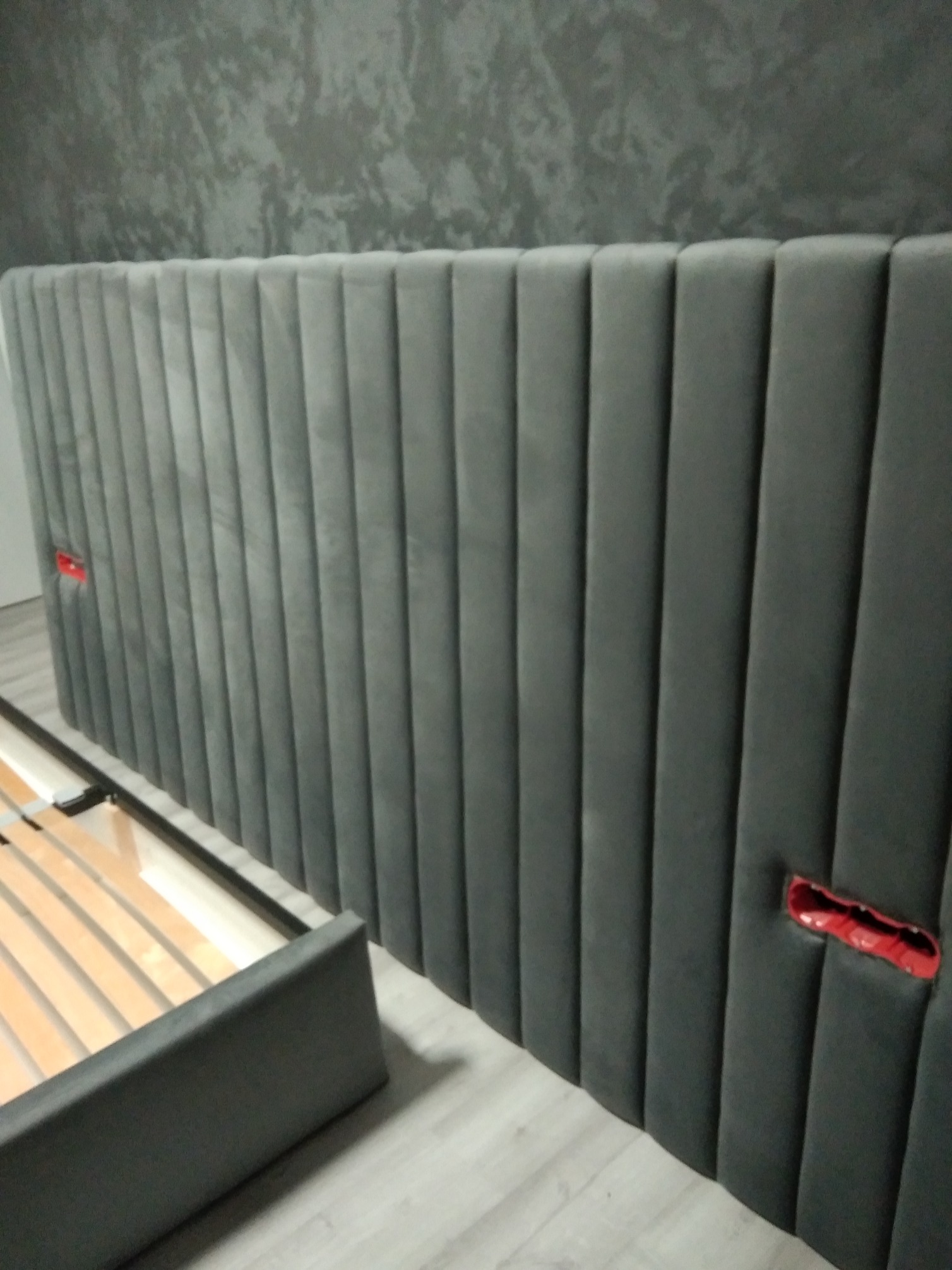 Szary, tapicerowany zagłówek łóżka z pionowymi przeszyciami, fragment ramy łóżka z drewnianym stelażem, ściana o fakturze betonu w tle.