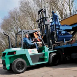 Zielony wózek widłowy Mitsubishi FD60-70 z operatorem, podnosi niebieski osprzęt na platformę ciężarówki.