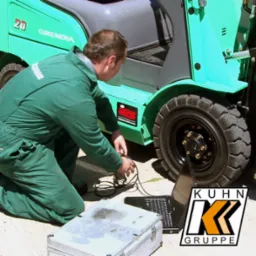 Serwisant w zielonym kombinezonie diagnozuje wózek widłowy Mitsubishi Grendia za pomocą laptopa i specjalistycznego sprzętu diagnostycznego firmy IPS, widoczne logo Kuhn Gruppe.