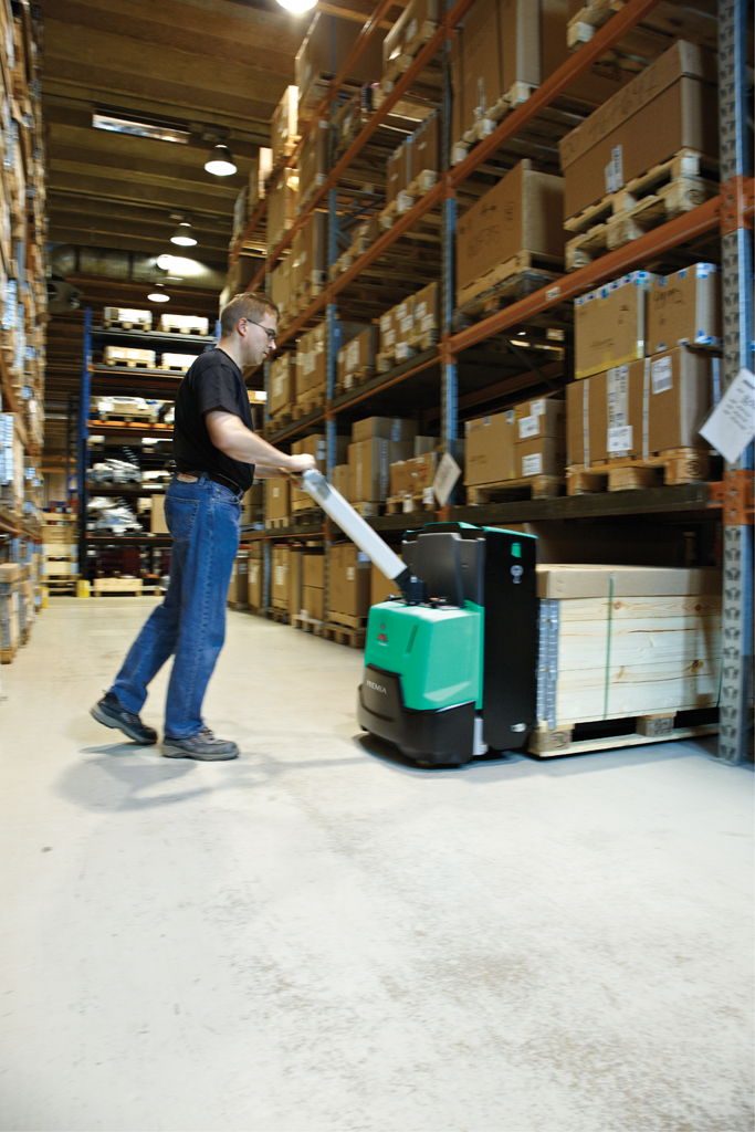 Pracownik magazynu obsługuje elektryczny wózek paletowy Mitsubishi PBP16-20N2, transportując paletę z drewnianymi elementami w głąb hali magazynowej z wysokim regałami wypełnionymi kartonami.