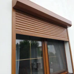 Brązowa roleta zewnętrzna częściowo opuszczona na oknie z moskitierą, widoczna biała elewacja budynku.