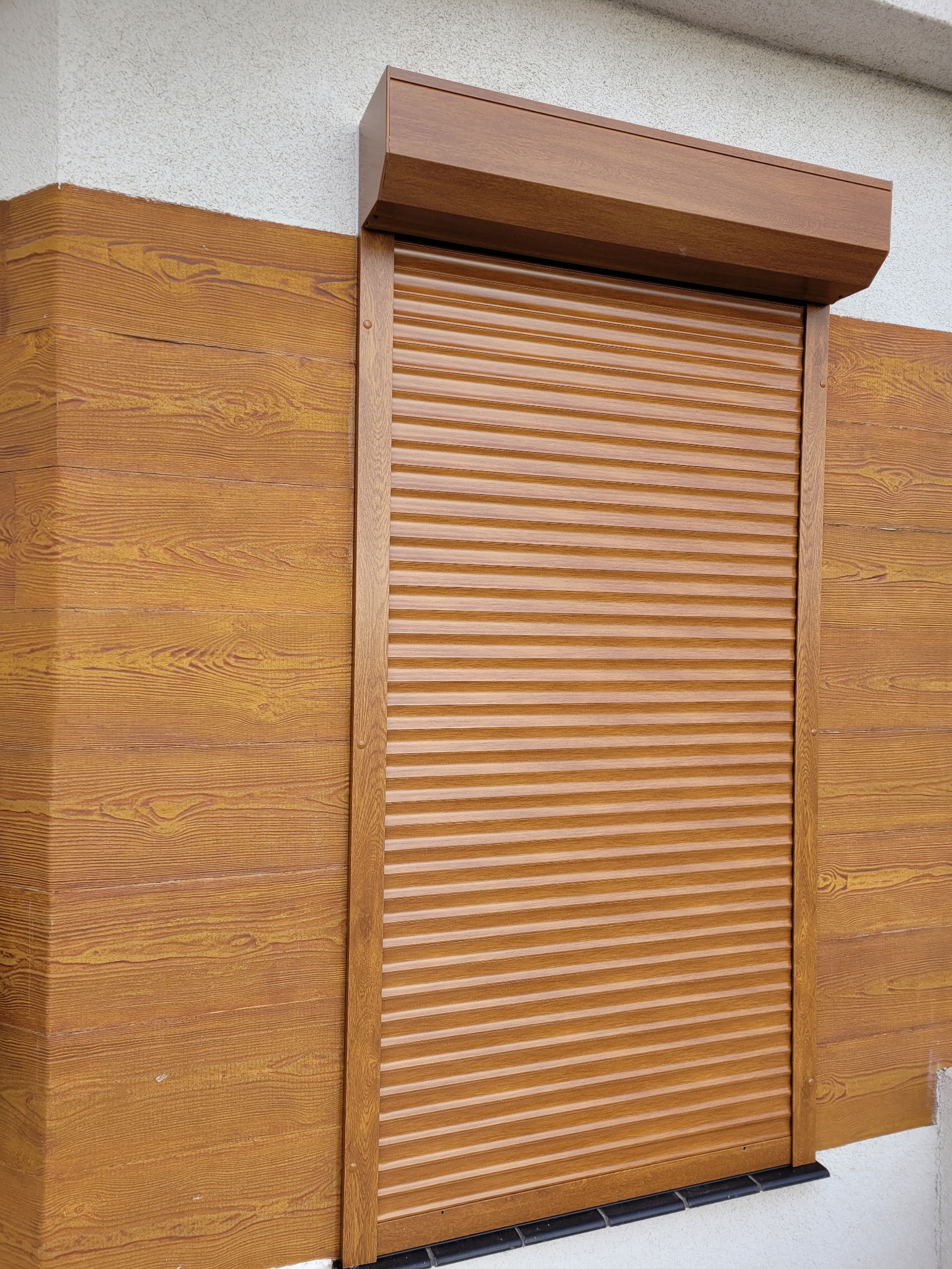 Brązowa roleta zewnętrzna, częściowo opuszczona, zamontowana na elewacji z imitacją drewna. Widoczne detale mocowania rolety.