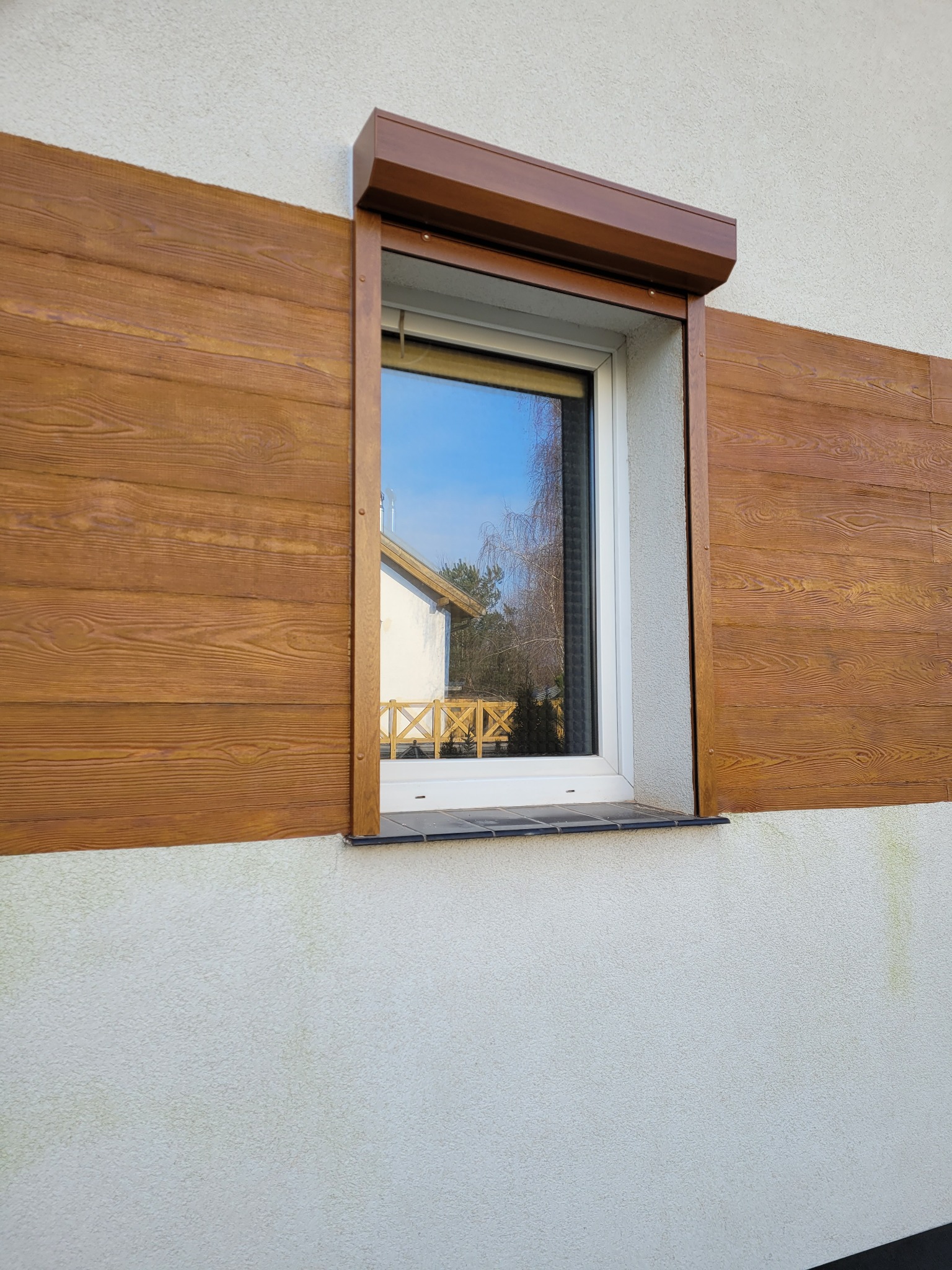 Brązowa roleta zwinięta nad oknem, obudowa imitująca drewno, widoczna faktura tynku na elewacji, odbicie nieba i drzew w szybie okna.