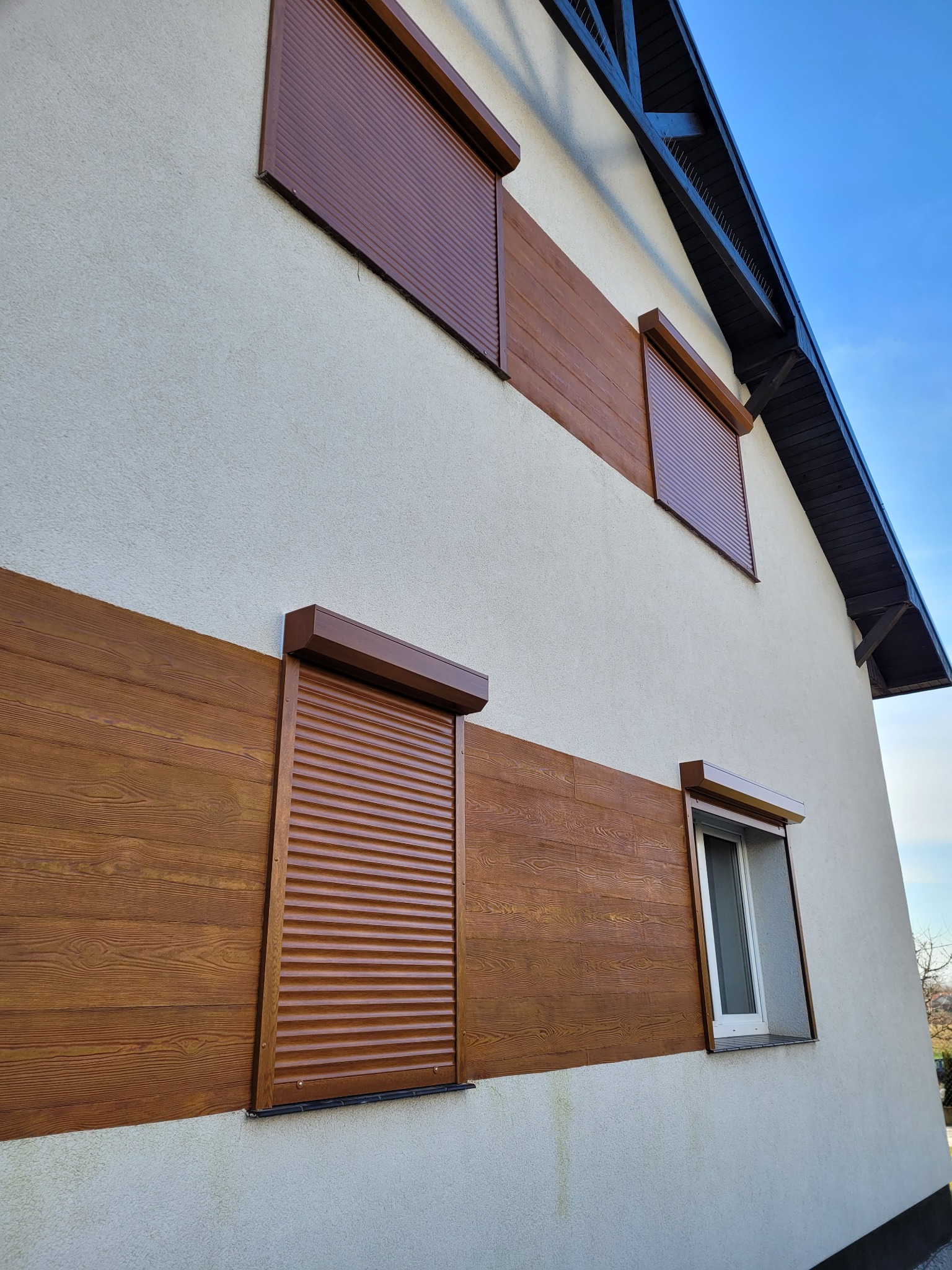 Brązowe rolety na elewacji domu z drewnianą okładziną, jedna roleta częściowo podniesiona, odsłaniająca okno z moskitierą.