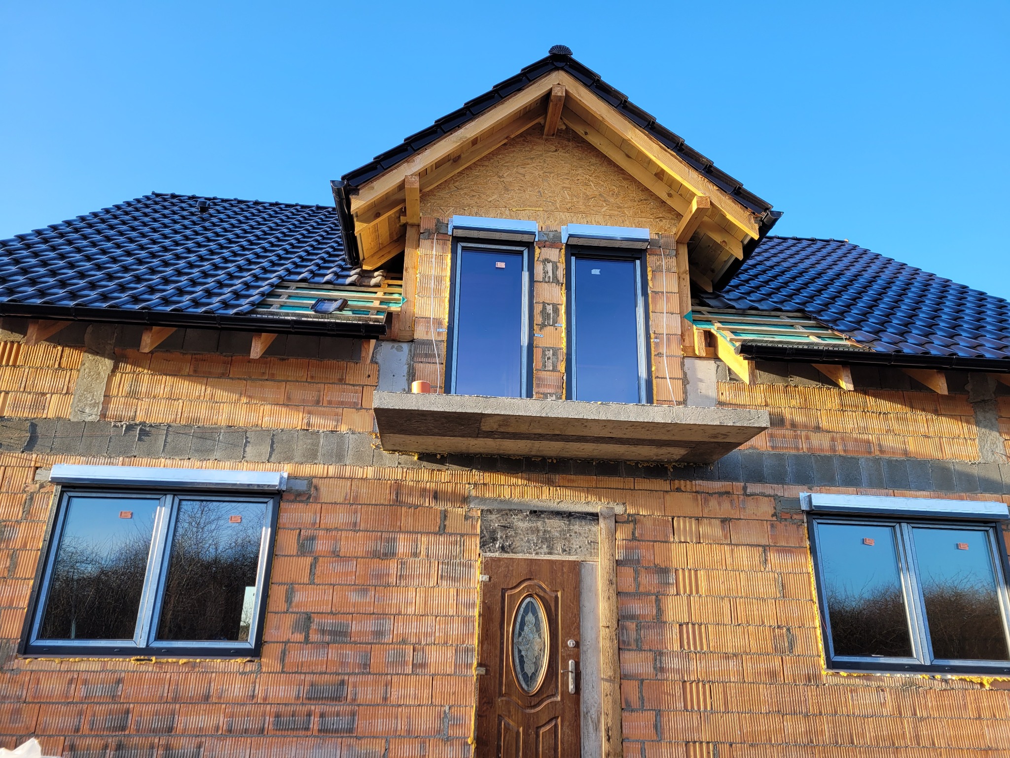 Surowy stan budynku z cegły z zamontowanymi roletami zewnętrznymi na oknach poddasza i parteru, widoczne elementy konstrukcji dachu i brakujące fragmenty elewacji.