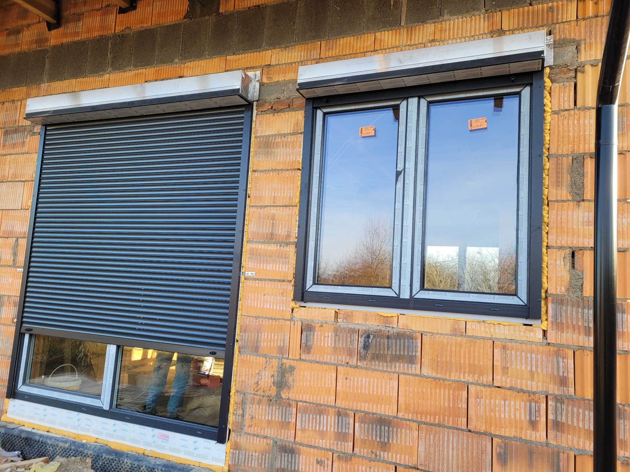 Częściowo opuszczona grafitowa osłona okienna na tle surowej cegły, obok okno z widocznym niebem i pianką montażową.