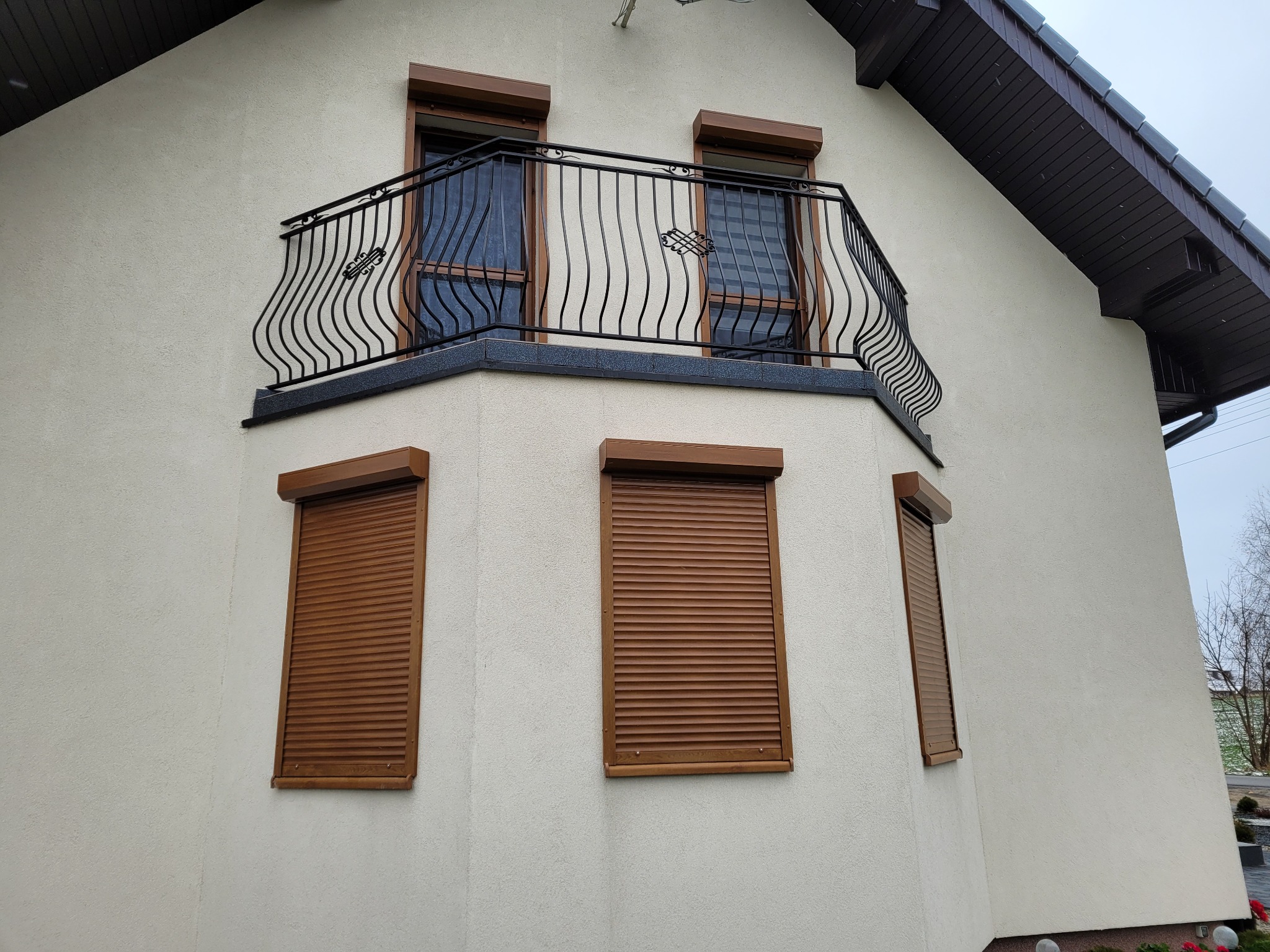 Elewacja domu z trzema oknami z brązowymi roletami zewnętrznymi oraz balkonem z czarną, ozdobną balustradą.