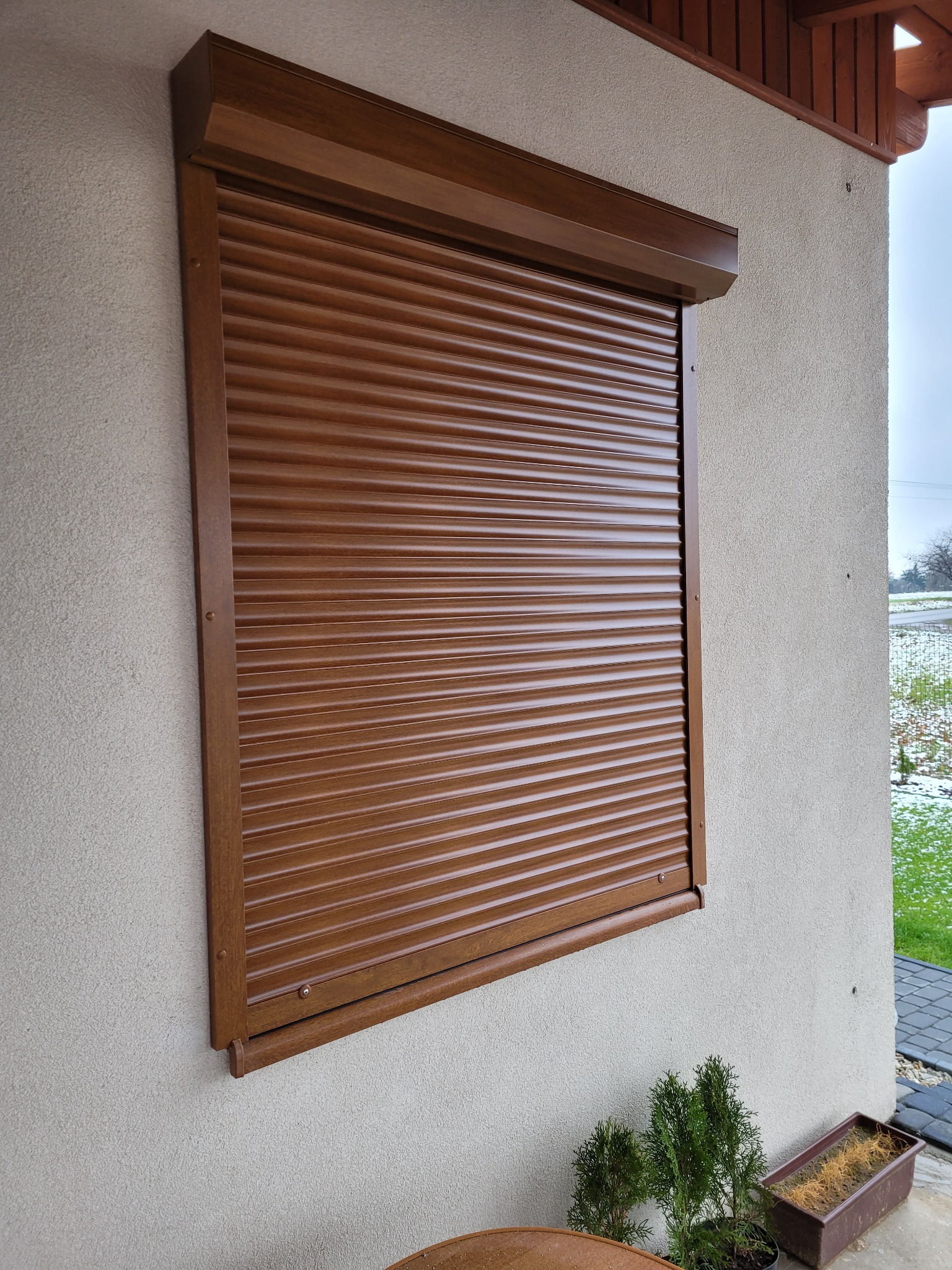 Zamknięta brązowa roleta zewnętrzna zamontowana na elewacji domu w słoneczny dzień, widoczne rośliny w doniczkach u dołu.