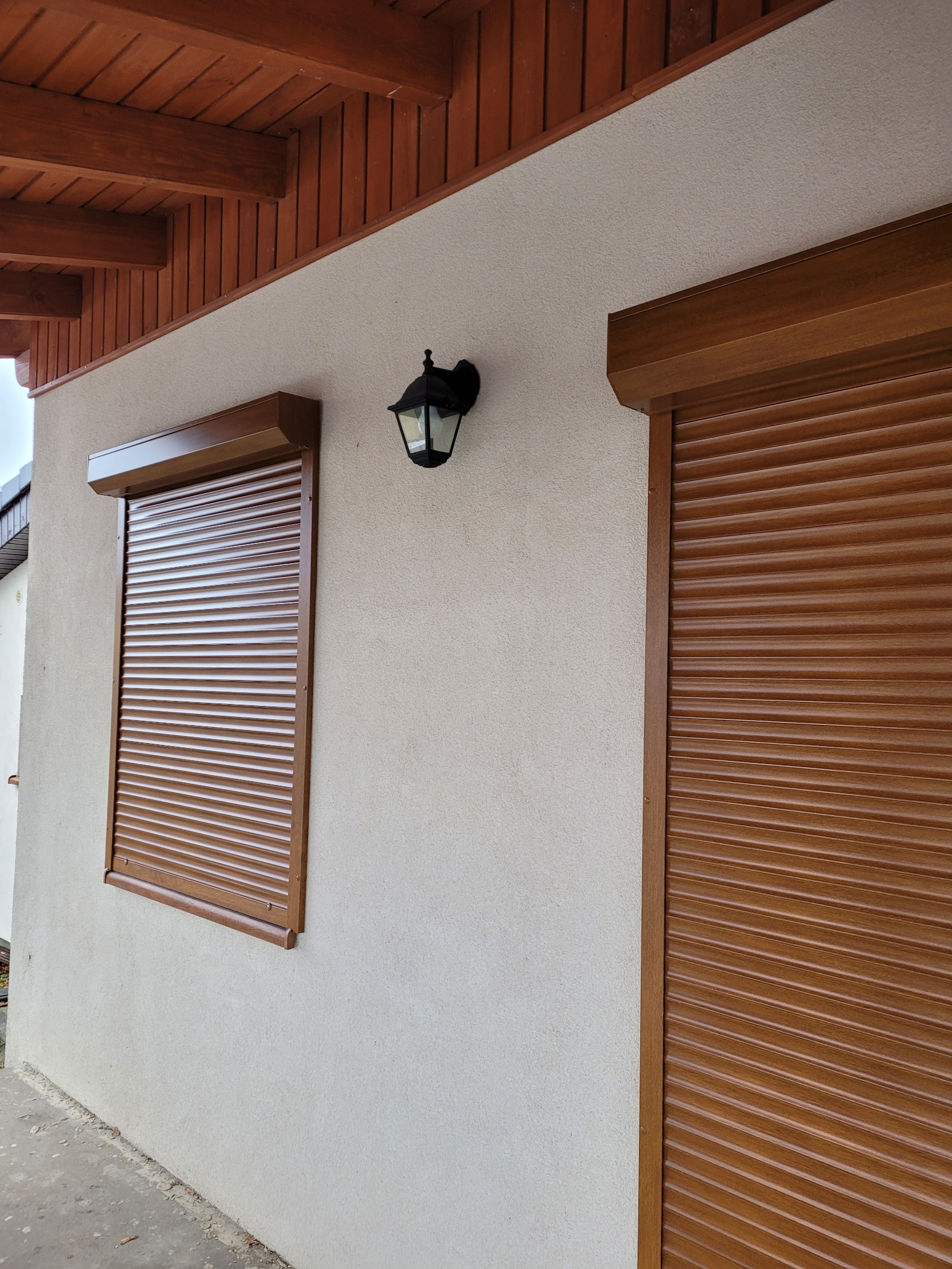 Brązowe rolety na oknach i drzwiach garażowych na tle beżowej elewacji domu z drewnianą podbitką dachową i czarną lampą zewnętrzną.