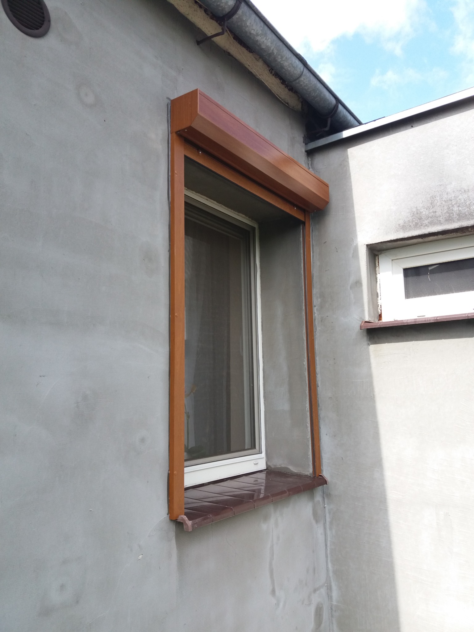 Zamknięta, brązowa roleta zewnętrzna zamontowana na oknie budynku z szarą, chropowatą elewacją, widoczny fragment rynny i małe okno obok.