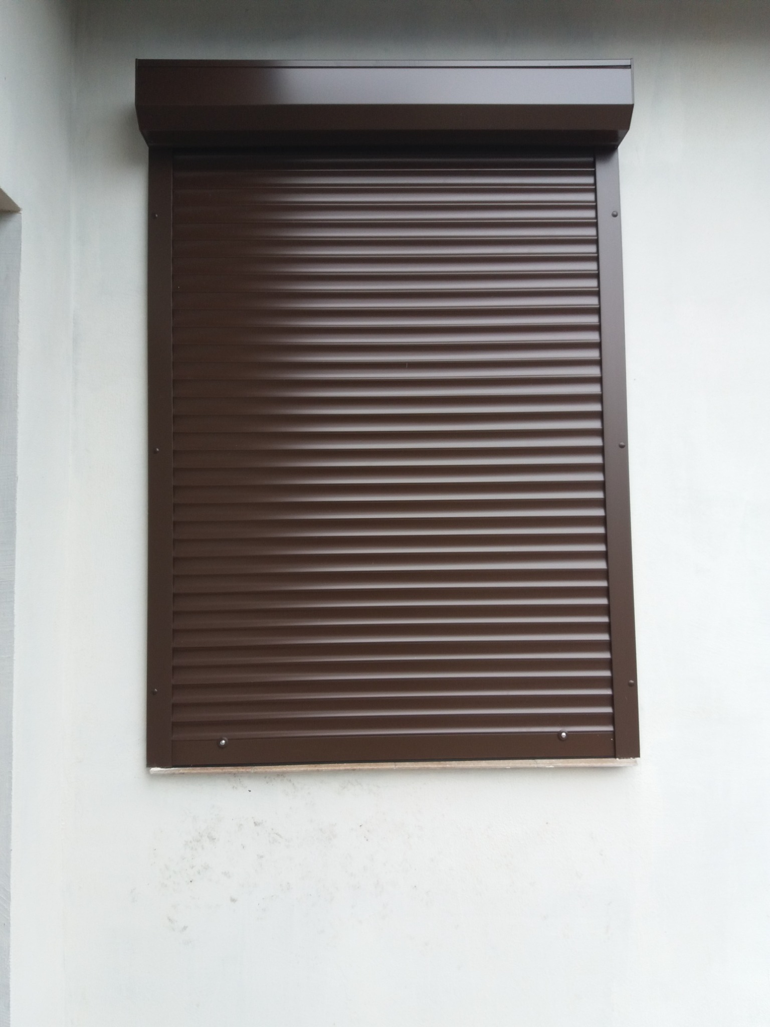 Zasłonięte, brązowe okno z roletą zewnętrzną zamontowaną na białej ścianie budynku.