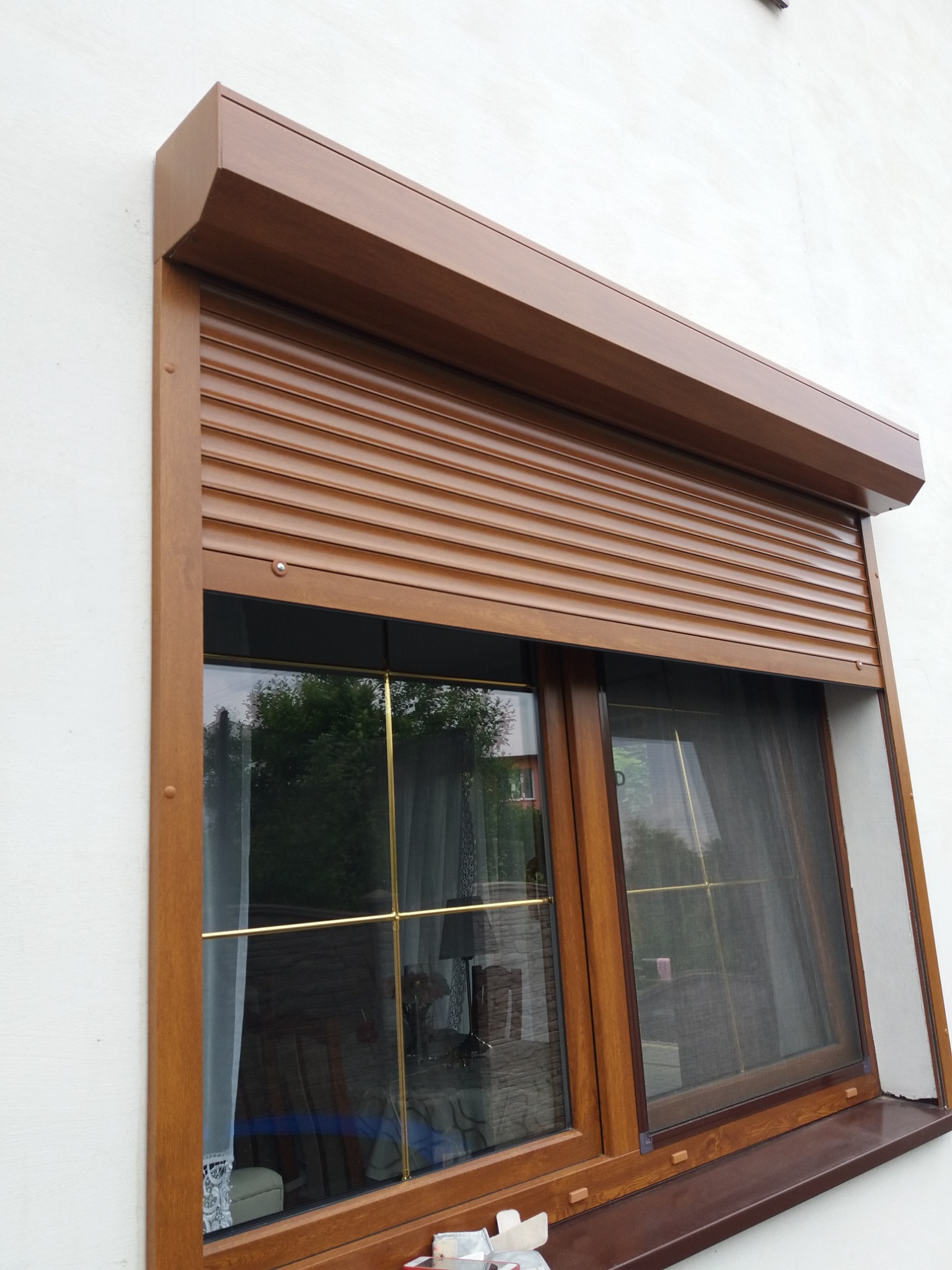 Brązowa roleta zewnętrzna częściowo opuszczona na oknie z moskitierą, widoczna biała elewacja budynku.