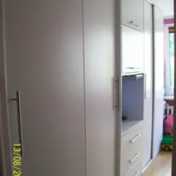 Biała szafa wnękowa z przesuwnymi drzwiami i chromowanymi uchwytami, widoczne szuflady i półki w otwartej wnęce, zabawki dziecięce w tle, zdjęcie z datą 13/08/2010.