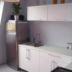 Kuchnia na poddaszu z białymi szafkami z prostymi uchwytami, stalową lodówką, zlewem oraz rośliną w srebrnym wiaderku na lodówce.