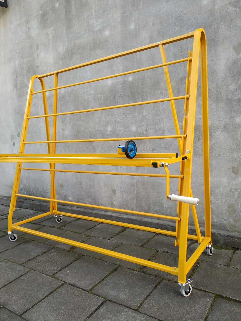 Żółty, metalowy stojak spawalniczy na kółkach z mechanizmem rolkowym, widoczny na tle szarej ściany i kostki brukowej.