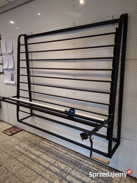 Czarna metalowa konstrukcja do cięcia materiałów z niebieskim pokrętłem, zamontowana na ścianie z perforacją.