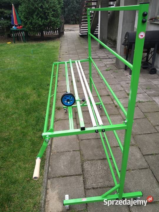 Zielony, metalowy stelaż na kółkach z korbą i niebieskim kołem, na zewnątrz, na tle trawnika i kostki brukowej.