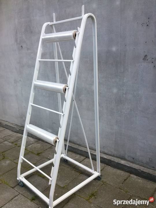 Biały, metalowy stojak na rolki folii stretch, z czterema rolkami na różnych poziomach, na kółkach, ustawiony na zewnątrz na tle szarej ściany.