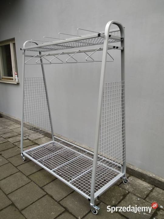 Metalowy, spawany wieszak na ubrania z siatkowymi półkami na kółkach, ustawiony na zewnątrz budynku z szarą elewacją.