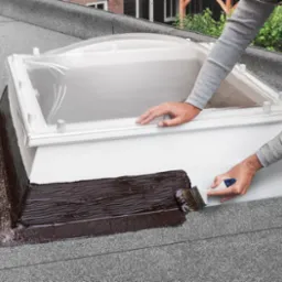 Uszczelnianie podstawy okna dachowego na płaskim dachu za pomocą pędzla i ciemnej masy bitumicznej, widoczny kominek wentylacyjny.