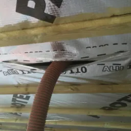 Poddasze w trakcie izolacji: widoczne drewniane belki, folia izolacyjna ALU 110 i czerwony wąż doprowadzający materiał izolacyjny.