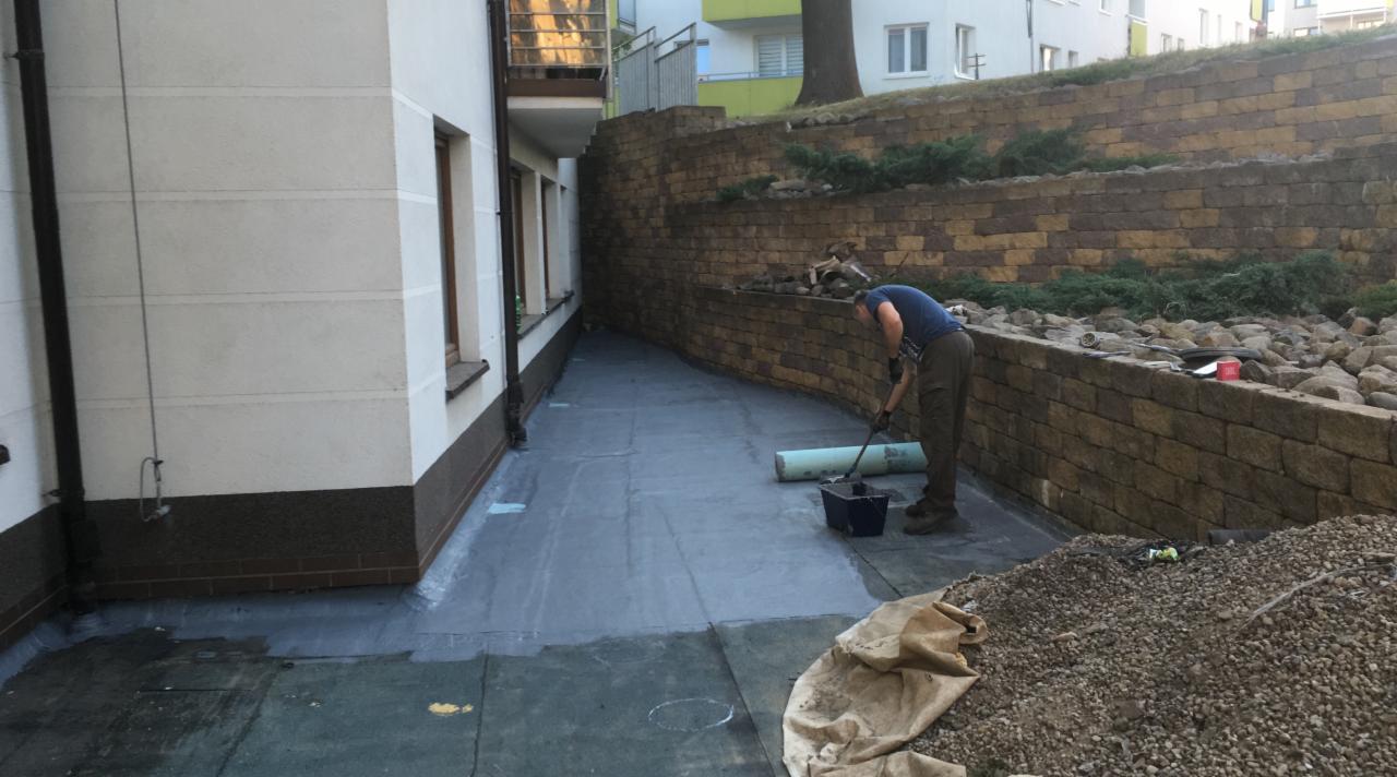 Pracownik z wałkiem malarskim rozprowadza izolację na płaskim dachu budynku mieszkalnego, widoczny fragment ściany budynku i mur oporowy z kamienia.