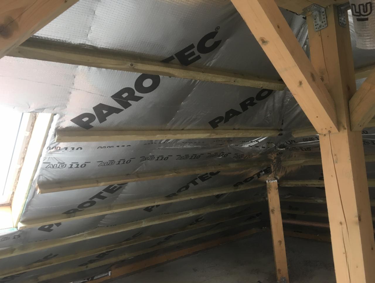 Izolacja poddasza folią paroizolacyjną Parotec ALU 110, widoczne krokwie i elementy konstrukcji drewnianej dachu, okno połaciowe w lewym górnym rogu.