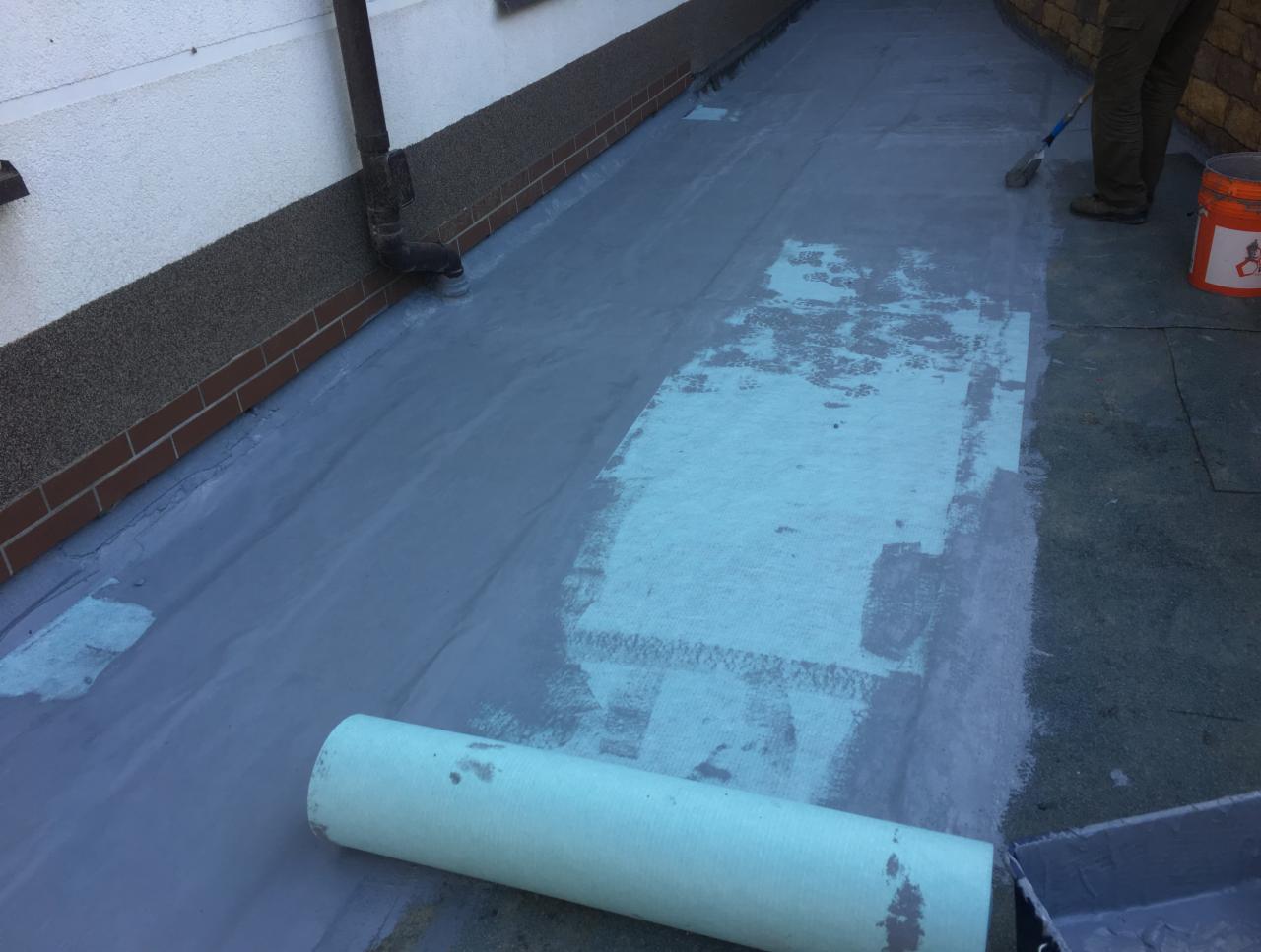 Aplikacja szarej hydroizolacji na płaskim dachu z widocznym wałkiem papy, pędzlem i wiadrem z farbą, na tle ściany budynku z rynną.