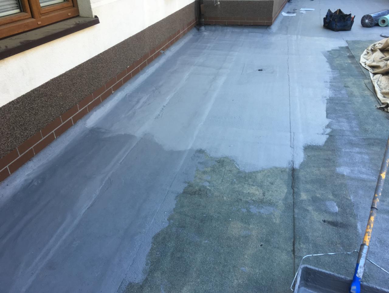 Płaski dach pokryty szarą, płynną membraną hydroizolacyjną, widoczny fragment ściany z ceglanym wykończeniem, narzędzia pracy i materiały w tle.