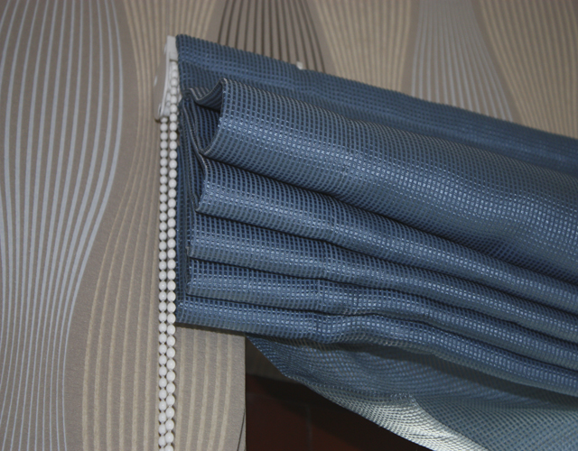 Niebieska roleta rzymska z delikatną fakturą w kratkę, zamontowana przy oknie z dekoracyjną tapetą w tle.