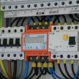 Rozdzielnica elektryczna z zamontowanym licznikiem energii, bezpiecznikami i okablowaniem. Widoczne elementy marki Eaton i Simlic. Szczegółowe ujęcie instalacji.