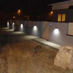 Nocna iluminacja posesji: rząd lamp ściennych oświetla ogrodzenie i wejście do domu. Widoczny fragment budynku z oknem i drzwiami, a także element krajobrazu - duży kamień.