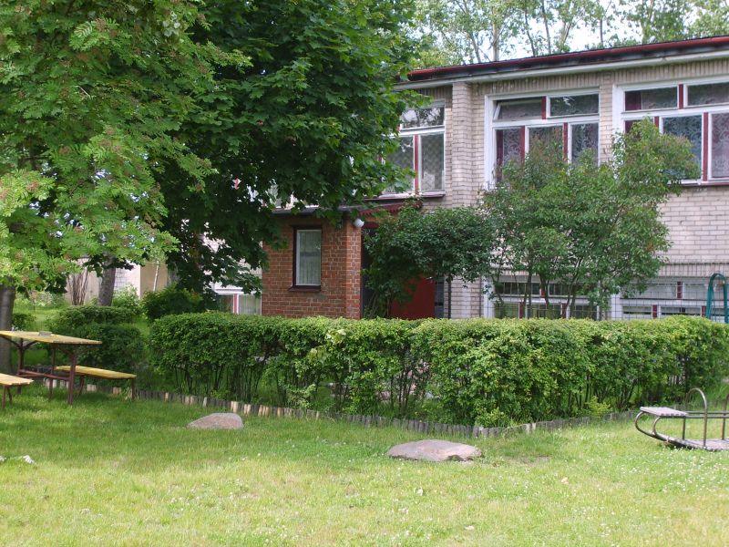 Budynek z czerwonymi akcentami okiennymi, otoczony zielenią, widoczny fragment placu zabaw i stolik piknikowy.