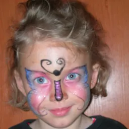 Uśmiechnięta dziewczynka z pomalowaną twarzą w motyle z brokatem, czarnym wzorem na czole i fioletowym nosem, na tle drewnianej ściany.