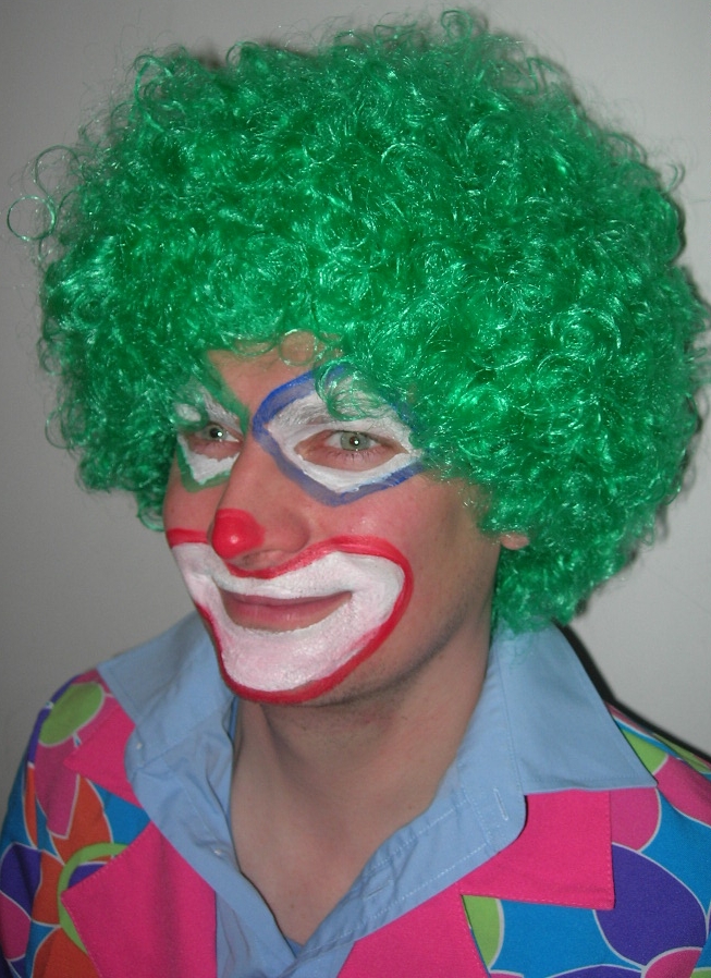 Portret klauna z zieloną peruką i pomalowaną twarzą, prezentującego się w Warszawie. Widoczny uśmiech i kolorowy strój.