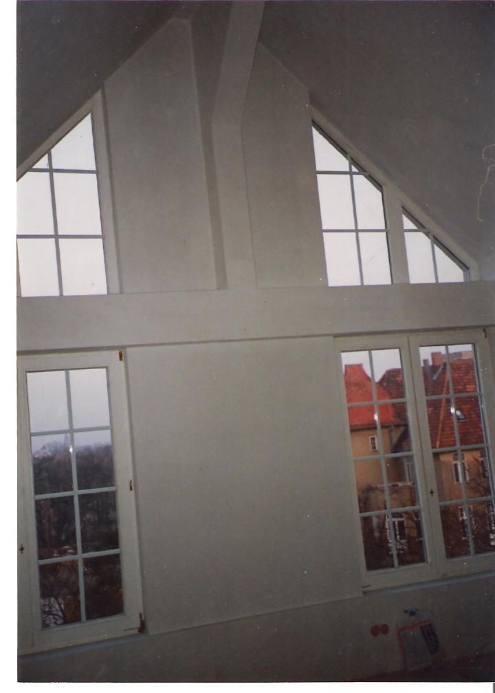 Wnętrze poddasza ze świeżo położonymi gładziami, widoczne okna trójkątne i prostokątne z widokiem na sąsiednie budynki.