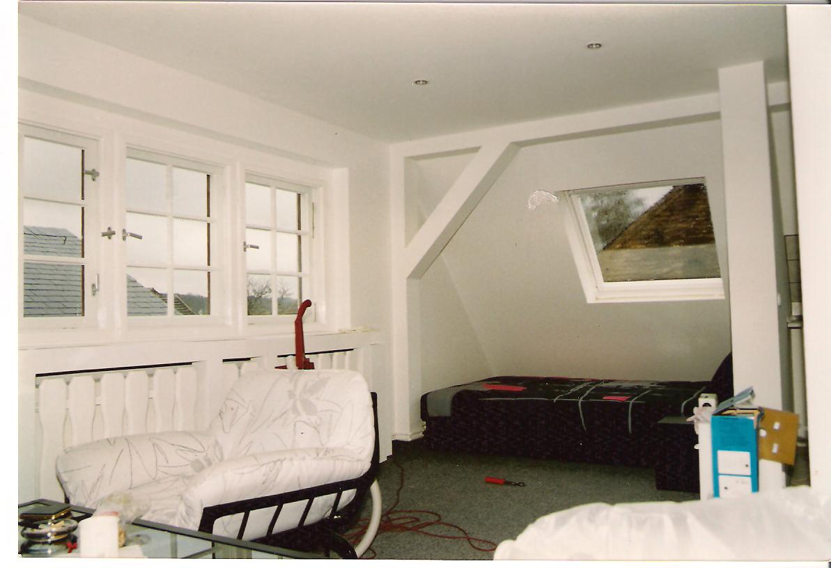 Poddasze po adaptacji: białe ściany, okna dachowe i pionowe, tapicerowany fotel z wzorem w kwiaty, widok na dach domu z zewnątrz.