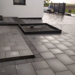 Nowoczesna, szara kostka brukowa ułożona wokół domu z minimalistycznym rabatem, widoczny fragment ogrodzenia panelowego i trawnik w tle.