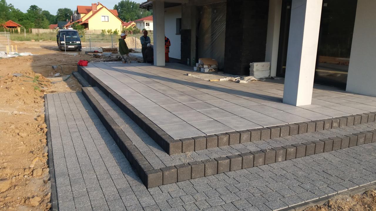 Nowoczesny taras z szarych płyt i ciemnych obrzeży, schody z kostki brukowej. W tle dom w trakcie budowy i pracownicy. Całość w jasnym, naturalnym oświetleniu.