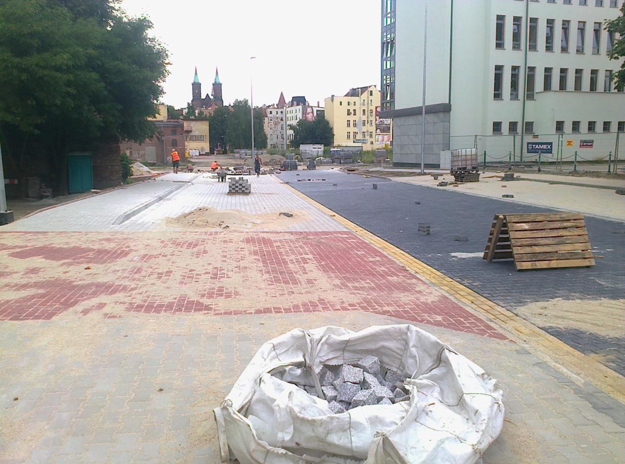 Ulica w trakcie budowy z częściowo ułożoną kostką brukową w kolorach szarości i czerwieni, workiem kostki na pierwszym planie i pracownikami w tle.