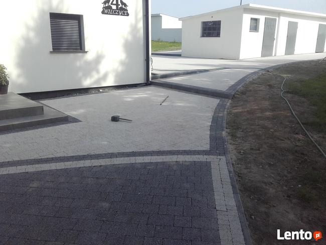 Ułożona kostka brukowa w Legnicy przed budynkiem z numerem 44. Widoczny fragment chodnika z ciemnoszarej kostki oraz jasnoszara powierzchnia wyłożona kostką z gumowym młotkiem leżącym na niej.