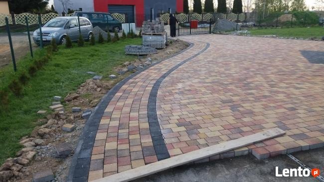 Układanie kostki brukowej na podjeździe z widocznymi paletami kostki i przygotowanym podłożem, częściowo zakończona realizacja.