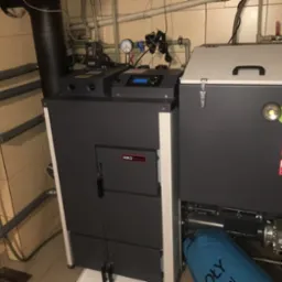 Nowoczesny piec na paliwo stałe Sokoł Max z certyfikatami Eco Design i klasy 5, zainstalowany w kotłowni z beżowymi płytkami i widocznymi rurami instalacji grzewczej.