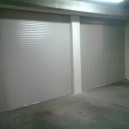 Dwie zamknięte, szare bramy rolowane w hali magazynowej, oznaczone słupami z żółto-czarnymi pasami ostrzegawczymi.